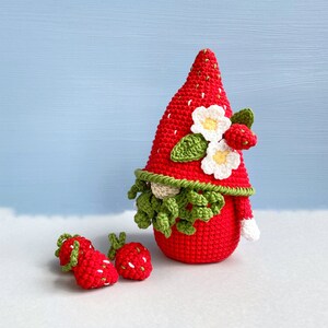 May include: A crocheted strawberry gnome with a red hat, white flowers, and green leaves. The gnome is standing on a white surface with two smaller crocheted strawberries.