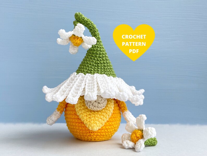 May include: A crocheted gnome with a green hat and white flower accents. The gnome is wearing a white crocheted flower hat and has a yellow and orange body. The gnome is sitting on a white surface. CROCHET PATTERN PDF.