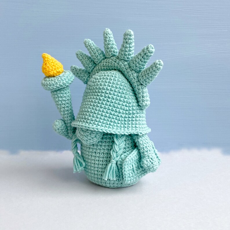 May include: A crocheted amigurumi statue of the Statue of Liberty, in a light blue color. The statue is holding a torch in its right hand.