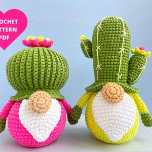Puede incluir: Dos gnomos cactus de crochet, uno con un corazón rosa y el otro con un corazón amarillo. Ambos gnomos tienen sombreros de cactus verdes y barbas blancas.