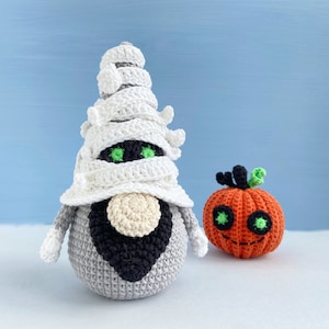 Pu&ograve; includere: Un gnomo mummia all'uncinetto con un cappello bianco a forma di benda e una barba nera, in piedi accanto a una zucca arancione all'uncinetto con occhi verdi e uno stelo.