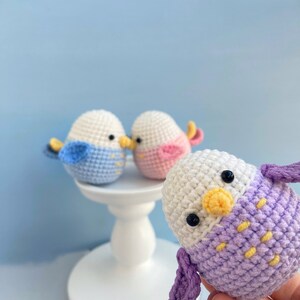 May include: Crocheted bird ornaments in white, blue, pink, and purple. The birds have black eyes and yellow beaks. Two birds are on a white pedestal, while a third is held in the hand. The background is a light blue.
