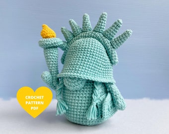Puede incluir: Patrón de crochet PDF para una muñeca amigurumi de la Estatua de la Libertad de color azul verdoso. La muñeca tiene una antorcha amarilla y una corona.