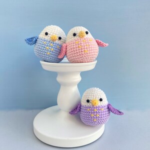 May include: Three handmade crochet parakeets in blue, pink, and purple, with white heads and yellow beaks. Two birds sit atop a white pedestal, while the third rests below. The birds have small wings and are set against a light blue background.