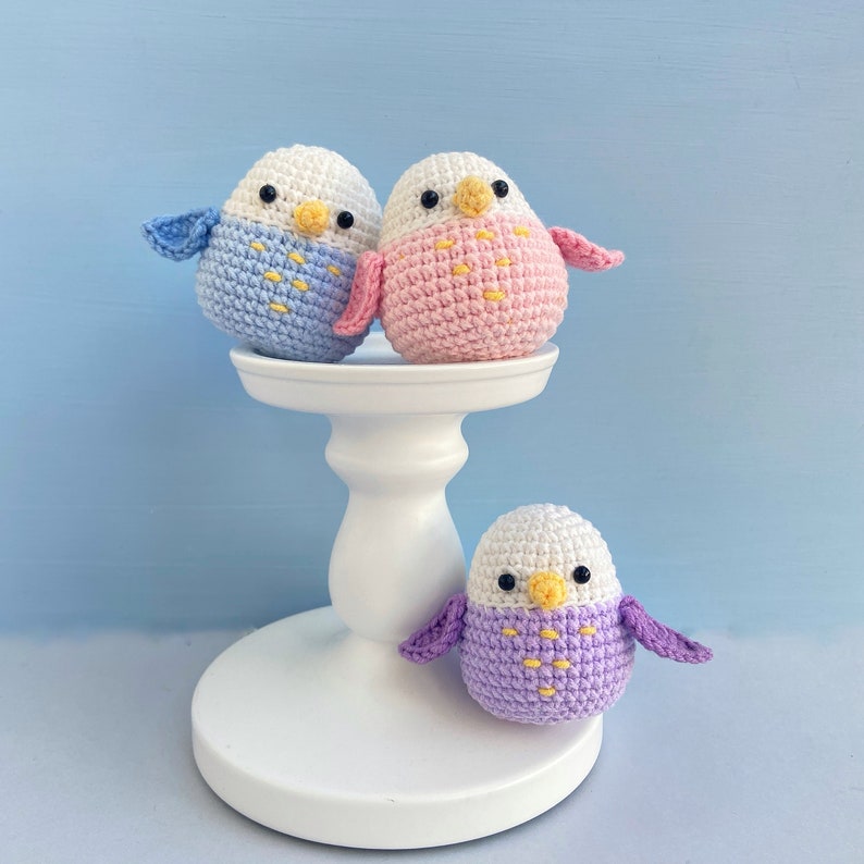 May include: Three handmade crochet parakeets in blue, pink, and purple, with white heads and yellow beaks. Two birds sit atop a white pedestal, while the third rests below. The birds have small wings and are set against a light blue background.