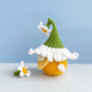 May include: A crocheted gnome with a green hat and white trim, holding a yellow flower. The gnome is sitting on a white surface.