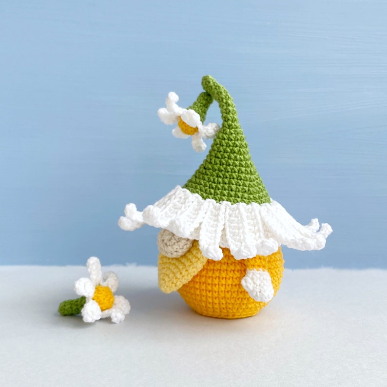 May include: A crocheted gnome with a green hat and white trim, holding a yellow flower. The gnome is sitting on a white surface.