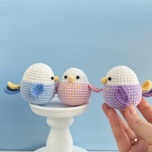 May include: Three handmade crochet bird ornaments in pastel colors. Each bird has a white head, a colored body, and small wings. One bird is blue, one is pink, and one is purple. They are displayed on a white pedestal against a light blue background.
