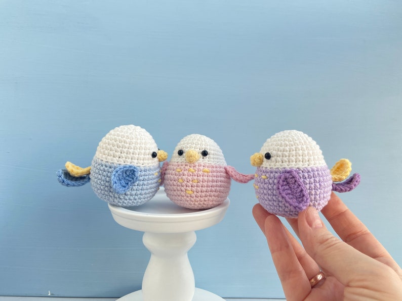 May include: Three handmade crochet bird ornaments in pastel colors. Each bird has a white head, a colored body, and small wings. One bird is blue, one is pink, and one is purple. They are displayed on a white pedestal against a light blue background.