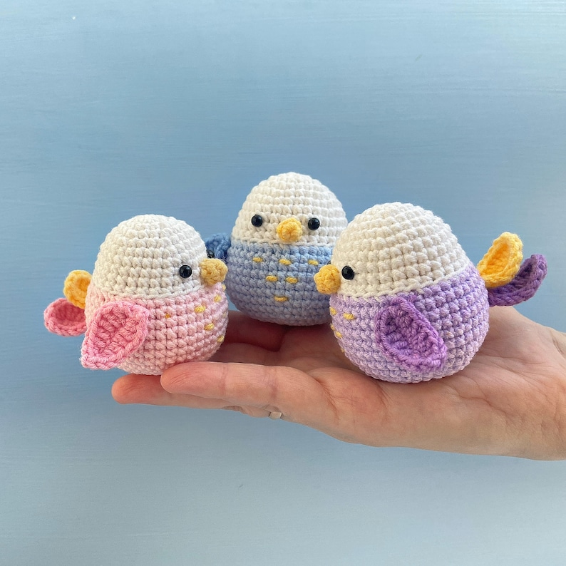 May include: Three handmade crochet birds in pastel colors. Each bird has a white head and a body in pink, blue, and purple. They have black eyes, yellow beaks, and colorful wings. The birds are small, measuring approximately 3 inches in height.