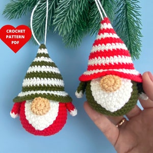 Op de afbeelding: Twee gehaakte gnome-versieringen, een met een groen-wit gestreepte hoed en de andere met een rood-wit gestreepte hoed. Beide gnomes hebben witte baarden en bruine neuzen.