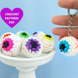 May include: Six crocheted eyeball keychains in various colors, including pink, purple, green, and blue. The keychains are displayed on a white cake stand. One keychain is being held by a hand, showing the silver chain and ring.