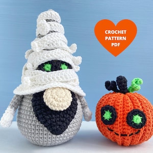Op de afbeelding: Een gehaakte mummie en pompoen Halloween-decoratie. De mummie is grijs en wit met een zwarte baard en groene ogen. De pompoen is oranje met groene ogen en een zwarte steel.