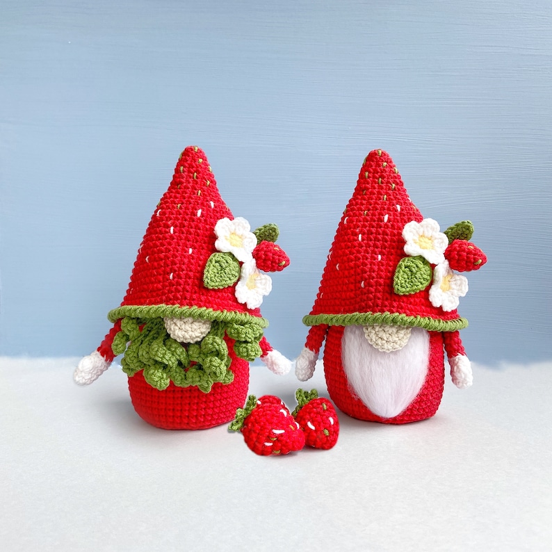 May include: Two crocheted strawberry gnomes with white flowers and green leaves. The gnomes are wearing red and white hats and have white beards. There are also four small crocheted strawberries in front of the gnomes.