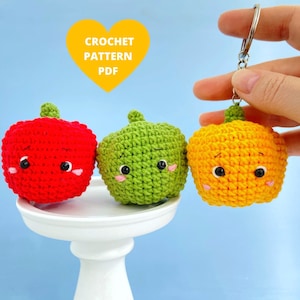 May include: Three crocheted vegetable keychains in red, green, and yellow. Each has a smiling face with black eyes and pink cheeks. The yellow keychain is attached to a silver keyring. The image includes the text "CROCHET PATTERN PDF".