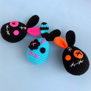 May include: Three crocheted bunny plushies with stitched-on eyes and mouths. The bunnies are black, blue, and orange.