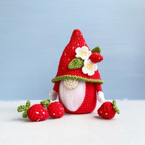 May include: A crocheted strawberry gnome with a white beard and a red and green hat with white flowers. There are three smaller crocheted strawberries in the foreground.
