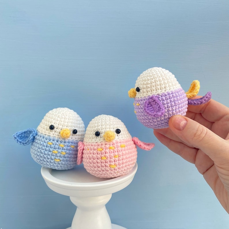 May include: Three handmade crochet birds in pastel colors. One bird is held by a hand. The birds have white heads, yellow beaks, and black eyes. One bird is blue, one is pink, and one is purple. The birds are on a white pedestal.