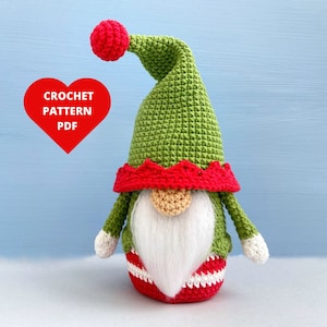 Puede incluir: Un gnomo de crochet verde con un gorro rojo y barba blanca. El gnomo está de pie sobre un fondo azul claro. El texto "CROCHET PATTERN PDF" está en un corazón rojo.