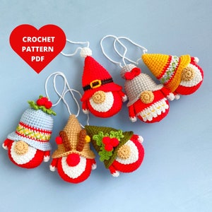 Könnte beinhalten: Sechs gehäkelte Gnom-Anhänger mit verschiedenen farbigen Hüten und Bärten. Die Anhänger sind rot, weiß, blau, grün, gelb und braun. Die Anhänger hängen an weißen Schnüren. Der Text "CROCHET PATTERN PDF" befindet sich in einem roten Herz.