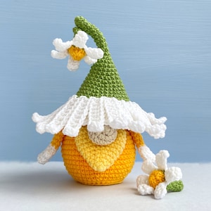 May include: A crocheted gnome with a green hat and white brim, a yellow and orange body, and a white daisy flower on top of the hat. There is a smaller white daisy flower with a green stem on the table next to the gnome.