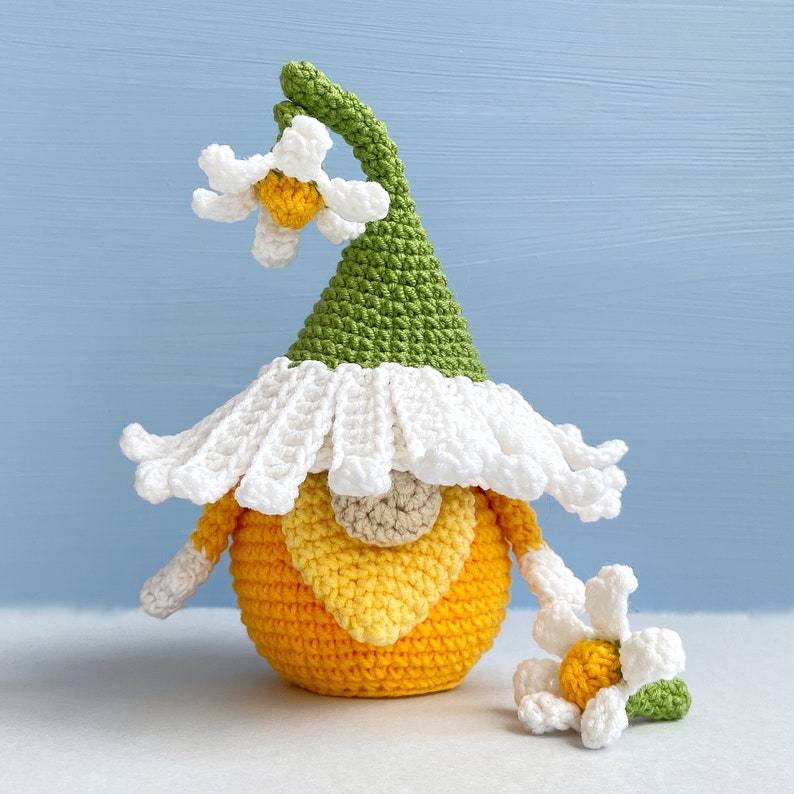 May include: A crocheted gnome with a green hat and white brim, a yellow and orange body, and a white daisy flower on top of the hat. There is a smaller white daisy flower with a green stem on the table next to the gnome.