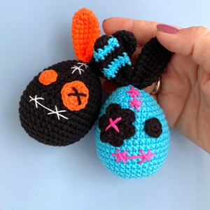 May include: Two crocheted plush toys shaped like skulls. One is black with orange and white details, the other is blue with black and pink details.