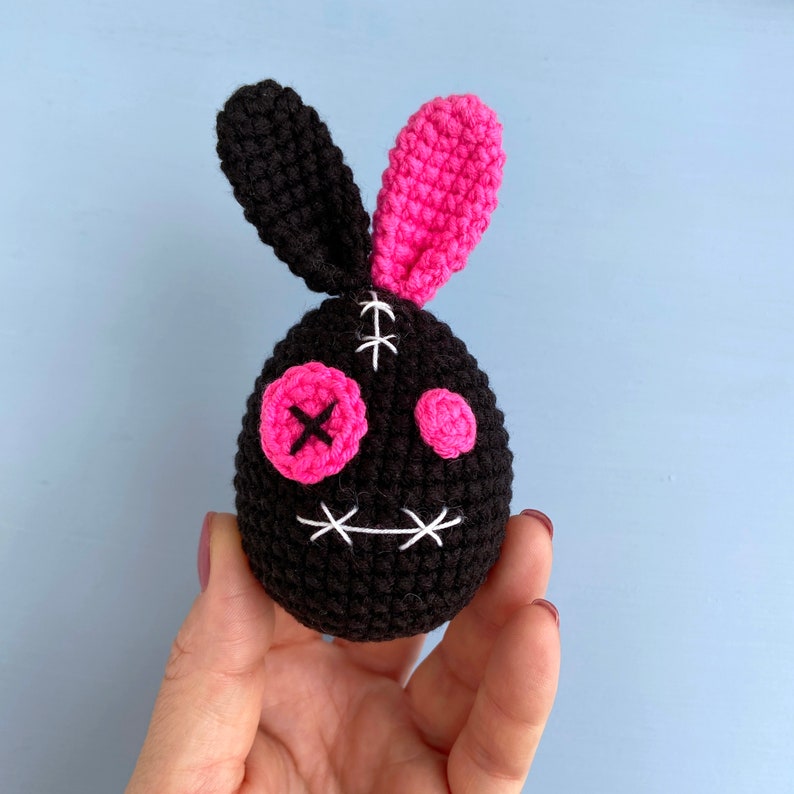 May include: A black and pink crocheted bunny with stitched eyes and a stitched mouth. The bunny has a stitched X on one eye and a pink button on the other eye.