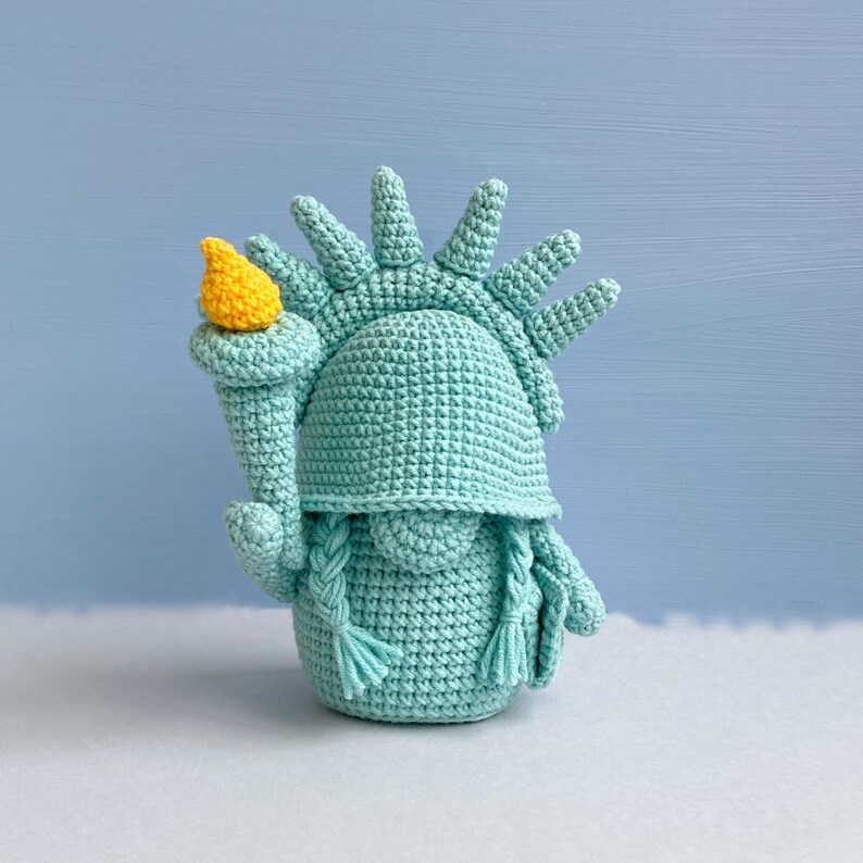 May include: A crocheted amigurumi statue of the Statue of Liberty, with a green body and a yellow torch.