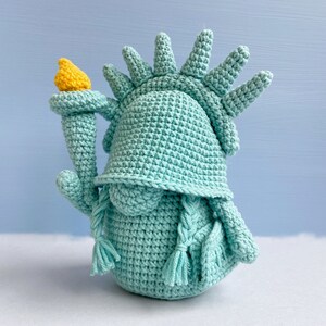 May include: A crocheted plush toy shaped like the Statue of Liberty. The toy is light blue and has a yellow torch.