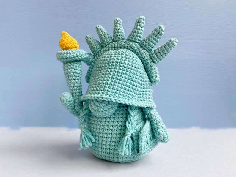 May include: A crocheted plush toy shaped like the Statue of Liberty. The toy is light blue and has a yellow torch.