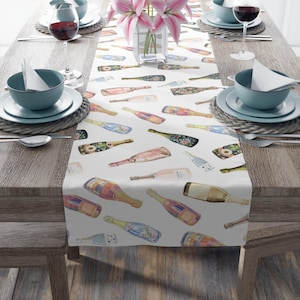 May include: White table runner with a repeating pattern of colorful champagne bottles. The bottles are illustrated in a watercolor style and are scattered across the runner.