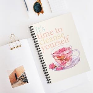 May include: A spiral bound notebook with a light beige cover featuring the text "It's time to cleanse yourself" in pastel colors. A glass teacup with pink rose petals floating in the tea is illustrated on the cover.
