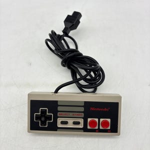 May include: A rectangular grey and black Nintendo video game controller featuring a black directional pad, two red circular buttons labeled B and A, and two grey oval buttons labeled SELECT and START, with a black cord and connector.