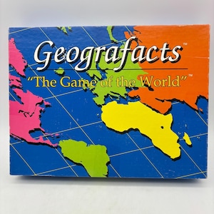 May include: A rectangular board game box titled Geografacts The Game of the World. The box features a colorful world map illustration with pink, green, yellow, and orange continents against a blue ocean background with thin yellow grid lines.