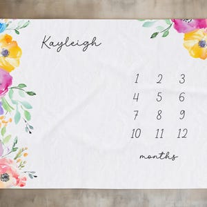May include: White baby milestone blanket with watercolour floral accents in pink, yellow, and blue. The blanket features the name "Kayleigh", numbers 1-12, and the word "months".