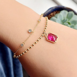 May include: A gold bracelet with two strands. One strand has small, round beads, and the other has small, clear gemstones. A rectangular, ruby-coloured gemstone pendant hangs from the beaded strand.