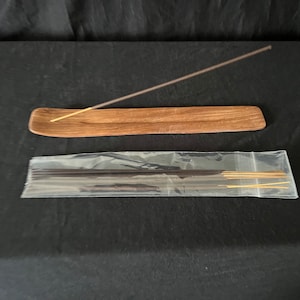 May include: A wooden incense holder with a dark brown finish and a single incense stick resting on top. A clear plastic bag containing a bundle of incense sticks is also shown.
