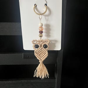 Puede incluir: Un llavero de macramé con forma de búho con un llavero dorado. El búho es beige con ojos negros y una borla. El llavero también presenta una cadena de cuentas en tonos blancos, marrones y dorados.
