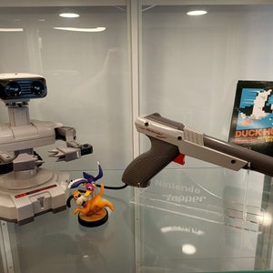 May include: A white and gray Nintendo Zapper light gun with a red trigger, a white and gray R.O.B. robot, and a Duck Hunt game cartridge. The Zapper is on a clear acrylic stand with the text "Nintendo Zapper" printed on it.