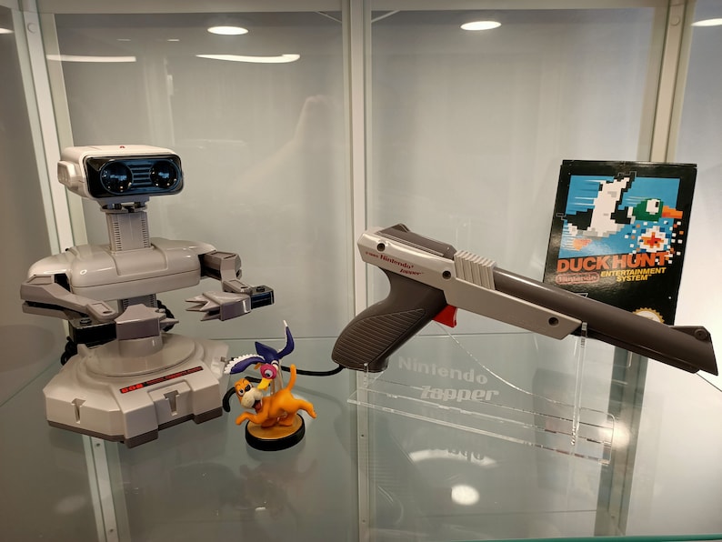 May include: A white and gray Nintendo Zapper light gun with a red trigger, a white and gray R.O.B. robot, and a Duck Hunt game cartridge. The Zapper is on a clear acrylic stand with the text "Nintendo Zapper" printed on it.
