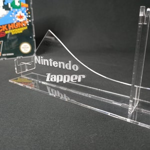 May include: A clear acrylic stand for a Nintendo Zapper light gun. The stand has a curved front and a back support with a slot for the Zapper. The stand is engraved with the words "Nintendo Zapper".