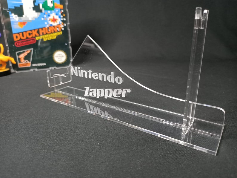May include: A clear acrylic stand for a Nintendo Zapper light gun. The stand has a curved front and a back support with a slot for the Zapper. The stand is engraved with the words "Nintendo Zapper".