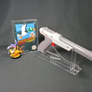 May include: A gray and white Nintendo Zapper light gun with a red trigger, displayed on a clear acrylic stand. The gun is labeled "Nintendo Zapper". A clear plastic case with a Nintendo Duck Hunt game cartridge is visible in the background.