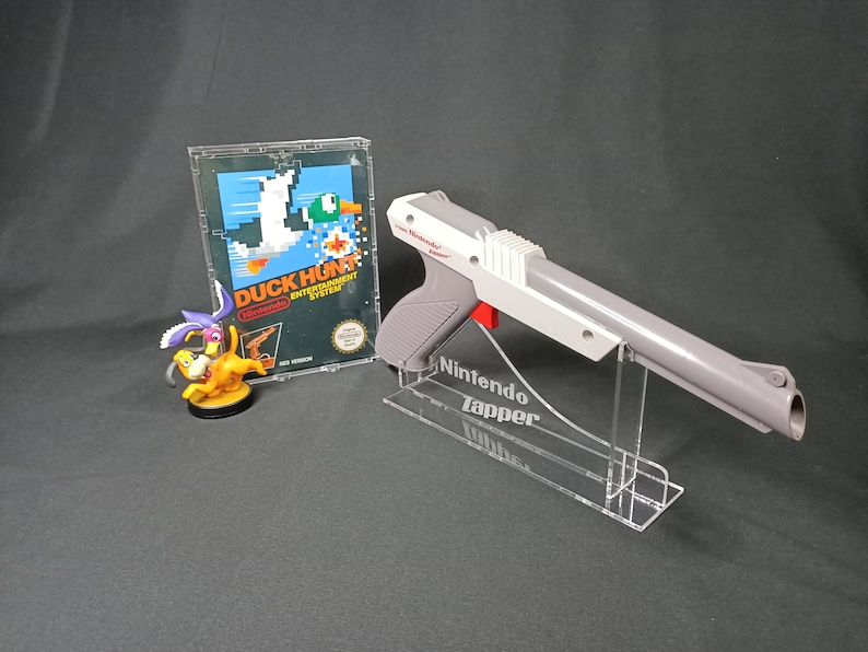 May include: A gray and white Nintendo Zapper light gun with a red trigger, displayed on a clear acrylic stand. The gun is labeled "Nintendo Zapper". A clear plastic case with a Nintendo Duck Hunt game cartridge is visible in the background.
