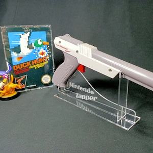 May include: A white and gray Nintendo Zapper light gun for the NES, with a red trigger, is displayed on a clear acrylic stand. The stand has the words "Nintendo Zapper" engraved on it. A Nintendo Entertainment System game cartridge for Duck Hunt is shown in the background.