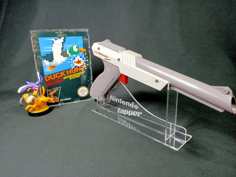 May include: A white and gray Nintendo Zapper light gun for the NES, with a red trigger, is displayed on a clear acrylic stand. The stand has the words "Nintendo Zapper" engraved on it. A Nintendo Entertainment System game cartridge for Duck Hunt is shown in the background.
