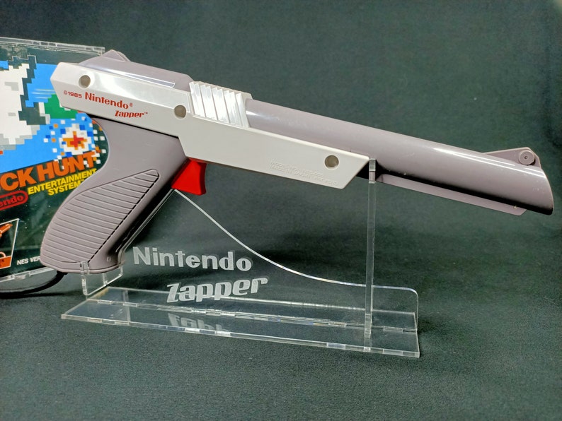 May include: A gray and white Nintendo Zapper light gun for the Nintendo Entertainment System. The Zapper is on a clear acrylic stand with the words "Nintendo Zapper" engraved on the stand.