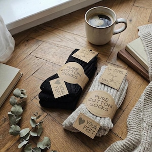 May include: Two pairs of ribbed socks, one black and one oatmeal-coloured, each wrapped in a brown paper band labelled PURE COTTON SOCKS and a small tag reading YOUR FEET with a heart icon. The socks are made of 100% cotton and feature a knit texture.