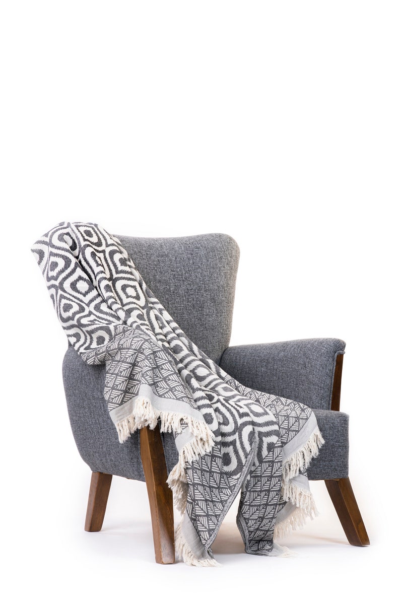 Puede incluir: Un sill&oacute;n gris con patas de madera y una manta de tiro con estampado en blanco y negro drapeada sobre el respaldo y el asiento.